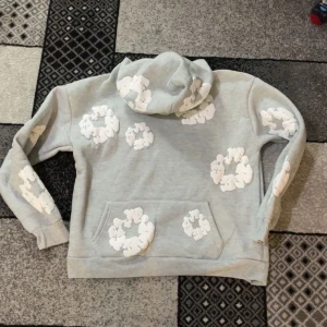 Grå hoodie med vita mönster från Denim Tears - Säljer en grå hoodie från Denim Tears med unika vita mönster över hela tröjan. Den har en klassisk känguruficka och en bekväm huva. Perfekt för en avslappnad stil.