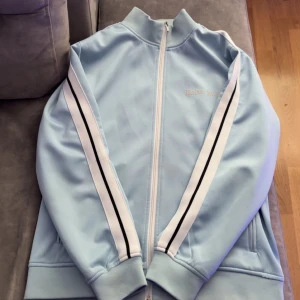 Ljusblå träningsjacka från Palm Angels - Säljer en ljusblå träningsjacka från Palm Angels med vita och svarta ränder längs ärmarna. Jackan har en dragkedja framtill och ribbade muddar vid ärmslut. Perfekt för en sportig look!