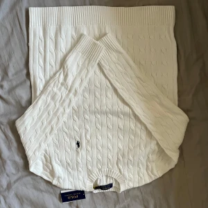Vit stickad tröja från Ralph Lauren - Säljer en stilren vit stickad tröja från Ralph Lauren med klassiskt kabelstickat mönster. Tröjan har en broderad logotyp i marinblått på bröstet och rund halsringning. Perfekt för en elegant och avslappnad look.