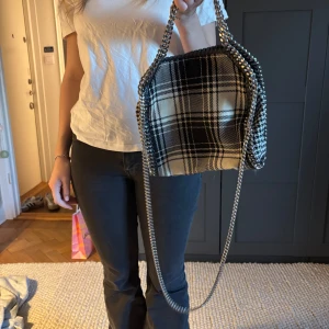 Stella McCartney Mini Falabella väska - Rutig axelväska med kedjedetaljer - Snygg rutig axelväska i svart och vitt med kedjedetaljer. Väskan har en unik design med ett rutigt mönster och en kedja som axelrem. Perfekt för att ge din outfit en trendig touch.