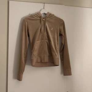 Beige hoodie från Juicy Couture - Säljer en snygg beige hoodie från Juicy Couture i mjukt sammetstyg. Den har en dragkedja framtill och en broderad logga på bröstet. Perfekt för en avslappnad stil med en touch av lyx. Huvan har justerbara snören med gulddetaljer. Aldrig använd💕