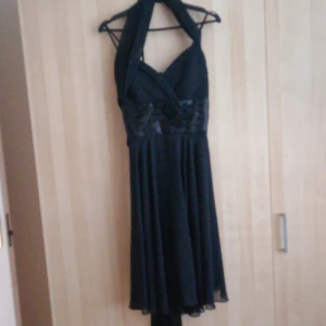 Svart klänning med halterneck - Elegant svart klänning med halterneck och vackra rosett-detaljer vid midjan. Klänningen har en omlott-design och är perfekt för festliga tillfällen.