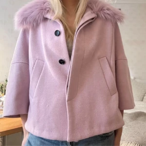 Ljusrosa kappa med fuskpäls - Säljer en elegant ljusrosa kappa med fuskpälsdetaljer på kragen. Jackan har trekvartsärmar och knäpps med stora knappar framtill. Perfekt för en stilren look.