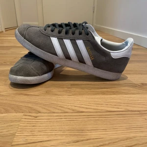 Adidas Gazelle sneakers - Snygga grå Adidas Gazelle sneakers i mocka med vita detaljer och klassiska tre ränder på sidan. Skorna har svart snörning och en vit sula.