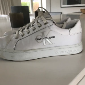 Calvin Klein vita sneakers Stl 38 - Ett par vita sneakers från Calvin Klein Jeans. Skorna har snörning och en stilren design med logotypen på sidan och baksidan. Stl 38, normal storlek. Äkta skinn o textil på ovansidan.  Använda ett fåtal gånger, inga defekter , är i bra skick. Kvitto finns. Nypris 1500:-