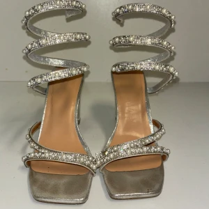 Silvriga klackskor med pärldetaljer - Eleganta silvriga klackskor med fyrkantig tå och transparent klack. Skorna har flera remmar dekorerade med pärlor och strass som slingrar sig upp runt vristen.
