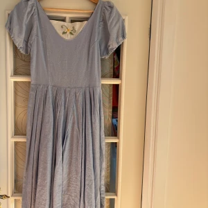 Vintage handgjord ljusblå klänning - Charmig handgjord ljusblå klänning med puffärmar och broderidetalj i urringningen. Klänningen har en knäppning i ryggen med dekorativa blomknappar. Perfekt för sommaren i ett linne-aktigt tyg. Köpt second hand men knappt använd efter det. (Av min lillasyster, klänningen är för liten för mig) - gissar xs.