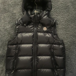 Moncler Bormes - Snygg svart dunväst från Moncler med dragkedja och huva. Västen har Moncler-logotypen på bröstet och praktiska fickor med dragkedja. Perfekt för våren.