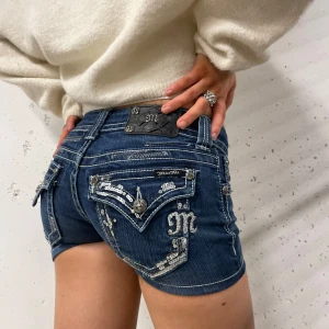 Jeansshorts från Miss Me - Snygga jeansshorts från Miss Me i mörkblå denim med dekorativa paljetter och broderier på bakfickorna. Shortsen har låg midja och en tight passform😊