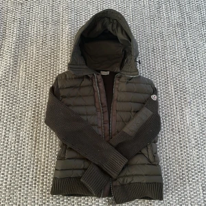 Mörkgrön dunjacka från Moncler - Säljer en snygg grön dunjacka från Moncler med stickade ärmar och huva. Jackan har dragkedja och två fickor framtill. Perfekt för sommarkvällarna med sin quiltade design och ribbade muddar. Säljer den då jackan är för liten nu. Men älskar den väldigt mycket och vill egentligen inte sälja. Scanner på märket funkar och den är inte trasig någon stans 