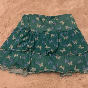 Grön kjol med fjärilsmönster från H&M - Söt grön kjol från H&M med ett fint fjärilsmönster i blått och vitt. Kjolen har volanger och är perfekt för en somrig look. Tillverkad i polyester vilket ger ett lätt och luftigt fall.