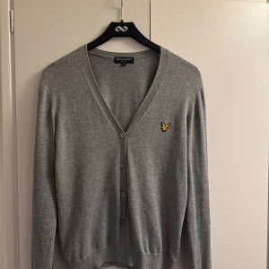 Grå kofta från Lyle & Scott - Säljer en stilren grå kofta från Lyle & Scott med v-ringning och knappar framtill. Den har ett broderat emblem på bröstet. Nedersta knappen har ramlat av. 