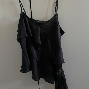 Svart ärmlös topp från Gina Tricot - Snygg svart ärmlös topp från Gina Tricot med volangdetaljer och justerbara axelband. Perfekt för en stilren look.