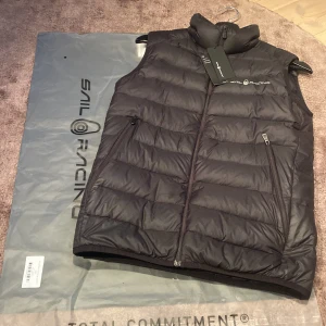 Svart dunväst från Sail Racing - Snygg svart dunväst från Sail Racing med dragkedja. påse och tags medföljer! Västen har en stilren design med logotyp på bröstet och praktiska fickor med dragkedja. Passar mig som är 173cm perfekt. Västen är fixad med lappar på 7 ställen typ där det är små hål men man tänker inte på att det är fixat om man inte kollar noga.                                                     Hör av er vid funderingar.                                             Kan fraktas📦