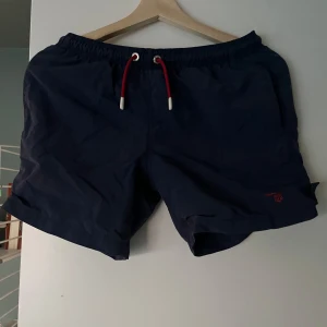Mörkblå badshorts från GANT - Snygga mörkblå badshorts från GANT med röd snörning i midjan. Perfekta för sommarens stranddagar. Tillverkade i ett lätt och bekvämt material.