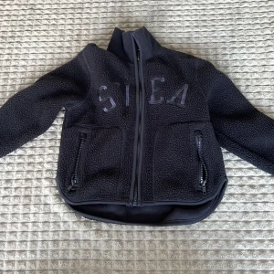 Svart teddyjacka från Svea - Säljer en svart teddyjacka från Svea med dragkedja framtill och två praktiska fickor med dragkedjor. Jackan har en hög krage och är perfekt för kyliga dagar. Den har en mjuk och fluffig textur som ger en mysig känsla.