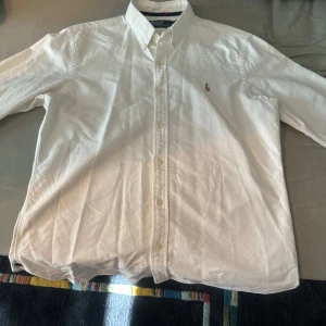 Vit skjorta från Ralph Lauren - Säljer en klassisk vit skjorta från Ralph Lauren med broderad logga på bröstet. Skjortan har långa ärmar och knappar framtill. Perfekt för en stilren look.
