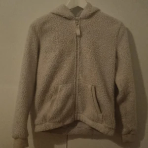 Beige teddyjacka med huva - Mysig beige teddyjacka med huva och dragkedja framtill. Jackan har två praktiska fickor och är perfekt för kyliga dagar. Den fluffiga texturen ger en trendig look.