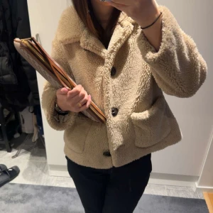 Beige teddyjacka  - Mysig beige teddyjacka med stora knappar framtill. Jackan har en avslappnad passform och två stora fickor. Perfekt för kyliga dagar.