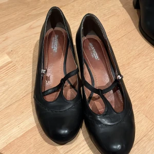 Svarta pumps med remmar - Snygga svarta pumps från Bianco Footwear med remmar över foten. Skorna har en låg klack och rundad tå, vilket ger en klassisk look. Perfekta för en stilren outfit.