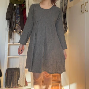 Grå smårandig klänning med knytning - Köpt secondhand, använd några få gånger. Säljer en stilren grå klänning med lång ärm och knytning framtill. Klänningen har en avslappnad passform och är perfekt för en enkel och elegant look.