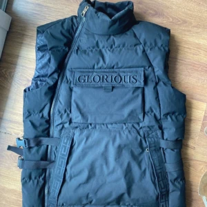 Svart dunväst från Glorious Gangsta - Säljer en svart dunväst från Glorious Gangsta med coola detaljer. Västen har en dragkedja framtill och justerbara spännen på sidorna. Perfekt för att hålla stilen när det är kyligt ute.