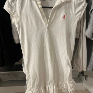 Vit klänning från Ralph Lauren - Säljer en vit klänning från Ralph Lauren med korta ärmar och en klassisk krage. Klänningen har en volangdetalj vid nederkanten och en broderad logotyp i rosa. Den har även en liten fläck som ej går bort.