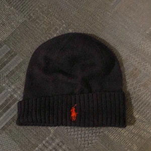 Svart mössa från Ralph Lauren - Snygg mörkblå mössa från Ralph Lauren med ribbad kant och ikonisk broderad logga i rött framtill. Perfekt för kyliga dagar.
