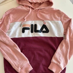 Rosa och vinröd hoodie från Fila - Säljer en snygg hoodie från Fila i rosa och vinrött med vit panel och logga på bröstet. Tröjan har justerbar huva med dragsko. Perfekt för en avslappnad stil!