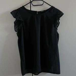 Svart blus med volang - Elegant svart blus med volangdetaljer vid ärmarna. Perfekt för en stilren look. Blusen har en rund halsringning och är tillverkad i ett lätt material.