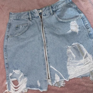 Ljusblå jeanskjol med dragkedja - Trendig ljusblå jeanskjol med slitna detaljer och råa kanter. Kjolen har en synlig dragkedja framtill, bälteshällor och klassiska fickor. Perfekt för dig som gillar en edgy och avslappnad stil.