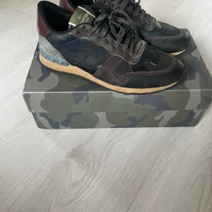 Valentino Rockrunner sneakers camouflage - Snygga Valentino Rockrunner sneakers med camomönster i svart, grått och mörkgrönt. Skorna har detaljer i mocka, skinn och textil samt vinröda hälpartier och nitar på sulan. Perfekta för dig som vill sticka ut med exklusiva och trendiga sneakers.