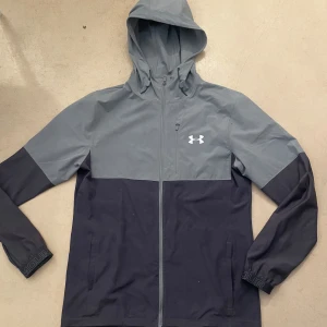 Grå och svart vindjacka från Under Armour - Snygg tvåfärgad vindjacka från Under Armour i grått och svart. Jackan har huva med dragsko, hel dragkedja framtill och praktiska fickor. Perfekt för dig som gillar sportig stil och vill ha något lätt och funktionellt använd 5 gånger max 