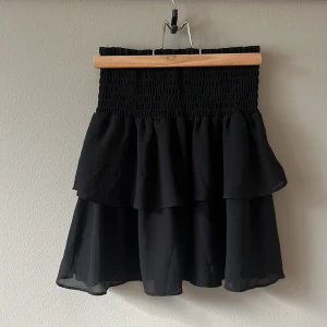 Svart volangkjol med smock - Supersöt svart kortkjol med smock i midjan och dubbla volanger. Kjolen har en luftig känsla och passar perfekt till sneakers eller boots. Enkel att styla med både crop top och stickad tröja.