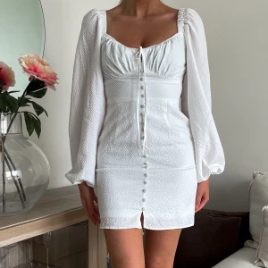 Vit kort klänning med puffärmar - Superfin vit kort klänning med puffiga långärmar, knappar framtill och söt knytning vid bysten. Klänningen har en lätt strukturerad yta och markerad midja som ger en snygg siluett.