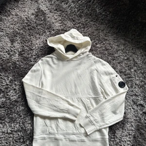 Vit hoodie från C.P. Company - Säljer en stilren vit hoodie från C.P. Company med klassisk huva och ficka framtill. Tröjan har lång ärm och den ikoniska linsen på vänster ärm. Tillverkad i mjuk bomull, perfekt för en clean och avslappnad look.