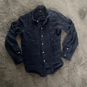 Mörkblå skjorta från Polo Ralph Lauren - Säljer en stilren mörkblå skjorta från Polo Ralph Lauren med klassisk krage och knappar. Skjortan har en broderad logotyp i rött på bröstet. Perfekt för en elegant look.