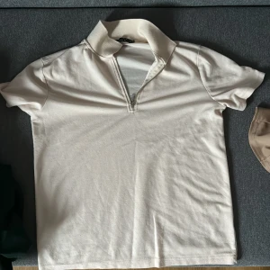 Beige pikétröja  - Snygg beige pikétröja från Delie i storlek M. Tröjan har korta ärmar, klassisk krage och en stilren half zip-dragkedja framtill. Perfekt för en clean och avslappnad look. Tillverkad i mjuk bomull som känns skön mot huden.