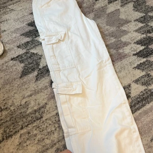 Vita cargopants med raka ben - Säljer ett par vita cargopants med raka ben och stora fickor på sidorna. Byxorna har normal passform, dragkedja och knapp framtill samt bälteshällor. Perfekta för en avslappnad och trendig look.