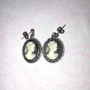 Vintage cameo örhängen i silverfärg - Snygga vintageinspirerade örhängen med oval form och klassiskt cameo-motiv i vitt mot svart bakgrund. Material rostfritt stål. Storlek :2,5 cm. Ramen är silverfärgad med detaljer runt om som ger en elegant och tidlös känsla. Perfekta för dig som gillar retrostil och unika accessoarer. 