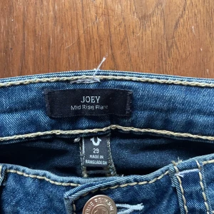 True Religion Joey Mid Rise Flare Jeans - Säljer ett par blå True Religion Joey jeans med midwaist och flare-ben. Klassiska kontrastsömmar, snygga fickdetaljer med knappar och logopatch bak. Jeansen är i bomull och har en riktigt schysst vintage vibe. Jag är 182 cm och dom sitter bra på mig i längden. Pris går att diskutera. 