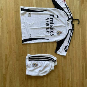 Real Madrid Bellingham matchtröja set - Säljer ett Real Madrid matchställ med Bellingham #5. Tröjan är vit med svarta detaljer, klubbmärke och sponsortryck. Korta ärmar och klassisk rund hals. Shortsen är vita med svarta ränder och klubbmärke. Tillverkad i lätt funktionsmaterial för bästa komfort.