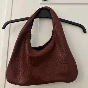 Brun/vinröd  axelväska i flätat skinn - Säljer en snygg brun/vinröd axelväska i flätat skinn. Väskan har en mjuk, rundad form och stängs med dragkedja upptill. Perfekt storlek för att få plats med allt du behöver och ger en cool vibe till din outfit.