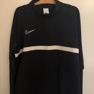 Svart Nike half zip tröja - Svart långärmad tröja från Nike med vit rand över bröstet och klassisk swoosh-logga. Tröjan har half zip-dragkedja och är gjord i ett sportigt, lätt material som passar perfekt till träning eller chill. Enkel och stilren design.