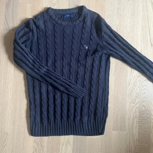 Mörkblå kabelstickad tröja från Gant - Kabelstickad mörkblå tröja från Gant med broderad logga på bröstet. Tröjan har rund halsringning, långa ärmar och ribbade muddar. Perfekt för dig som gillar klassisk och stilren look. Tröjan har en ganska tight passform och passar nog bra för någon som är runt ~160cm lång. Priset inte hugget i sten!!!