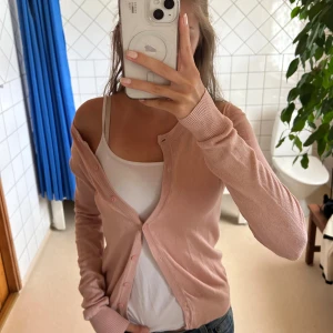 Ljusrosa kofta med knappar - Söt ljusrosa kofta med rund hals och knäppning framtill. Långärmad modell i tunt, mjukt material som sitter snyggt och är lätt att styla över ett linne eller t-shirt. Perfekt för lager-på-lager och passar till både jeans och kjol.