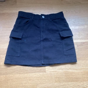 Svart kortkjol med cargofickor H&M - Svart kortkjol från H&M med cargofickor på sidorna och klassisk jeansknapp framtill. Kjolen har bälteshällor och är gjord i ett stadigt bomullstyg. Perfekt för en chill och trendig look.