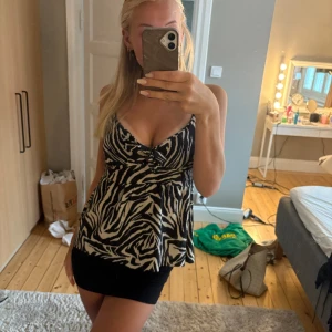 Zebramönstrad linne-topp med rosett - Snygg linne-topp med svartvitt zebramönster och smala axelband. Toppen har en söt rosettdetalj framtill och lätt utställd passform som ger en flowy känsla. Perfekt för dig som vill sticka ut med ett djurmönster. Materialet känns lätt och luftigt.