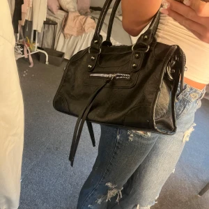 svart väska  - Snygg svart handväska i skinn med coola detaljer som silverfärgad dragkedja framtill och långa remmar. Väskan har två handtag och rymligt fack med dragkedja upptill. Perfekt för dig som gillar en edgy och stilren look.
