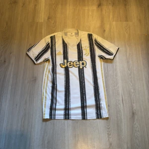 Juventus hemmatröja Adidas 2020/21 - Juventus hemmatröja från säsongen 2020/21 med klassiska svarta ränder på vit botten, gulddetaljer och Jeep-logga på bröstet. Tröjan har korta ärmar, V-ringning och är tillverkad i lätt och ventilerande material. Perfekt för dig som älskar fotboll och vill visa ditt stöd för Juve.
